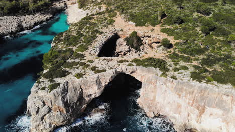 a-drone-shot-flying-backwards-away-from-a-sea-arch-with-a-couple-of-tourists-entering-the-frame-near-cala-varques-in-majorca