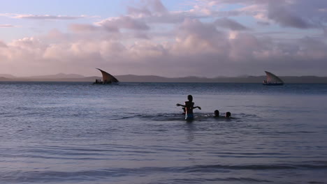 Kinder-Genießen-Es,-Im-Meer-Zu-Schwimmen,-Während-Im-Hintergrund-Ein-Traditionelles-Pirogenboot-Bei-Sonnenuntergang-Segelt