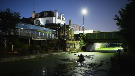 Man-kayaking-on-green-oozed-canal-with-surrounded-by-houses-at-night,-wide-shot