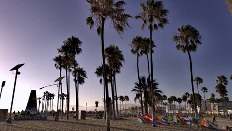 Palm-trees-waving-in-a-gentle-breeze,-while-people-enjoy-a-warm-sunset-at-the-Venice-beach-Boardwalk,-in-Los-Angeles,-California,-USA---Handheld-shot