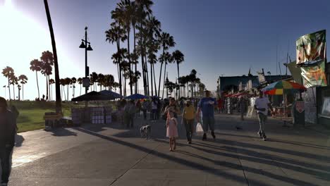 People-walking-by-and-around-the-Venice-Boardwalk-wearing-masks,-during-golden-hour,-in-Los-Angeles-during-Covid-19-pandemic---handheld-static