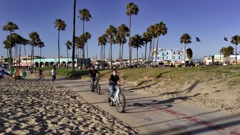 People-riding-bikes-along-Venice-Beach-Boardwalk-during-sunset-golden-hour,-in-Los-Angeles,-Califonia,-USA