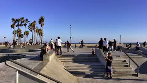 People-skating-and-walking-around-at-the-Venice-Beach-Skate-Park-with-ocean-and-mountains-in-background,-on-a-sunny-day,-in-Los-Angeles,-California,-USA---handheld-shot