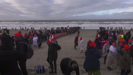 Red-cap-wearing-Dutch-people-getting-ready-to-celebrate-New-Year's-dive-at-Texel-Beach-in-North-Holland,-Netherlands---Wide-static-shot