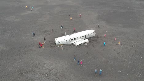 Drone-flying-over-famous-Plane-wreck-in-iceland-with-black-sand-and-mountains-in-the-background,-lot-of-tourist-walk-around-the-attraction
