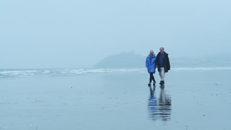 Elderly-couple-holding-hands,-walking-and-enjoying-stroll-on-Criccieth-Beach-on-misty-day-in-stormy-and-windy-weather-in-Wales