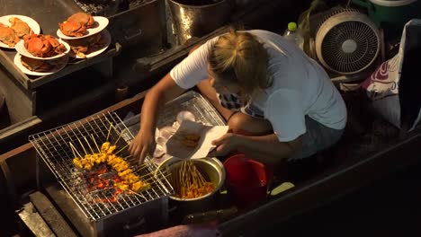 Thai-Woman-Preparing-Fried-Meat-Sticks-on-Boat-at-Traditional-Floating-Night-Market,-Thailand