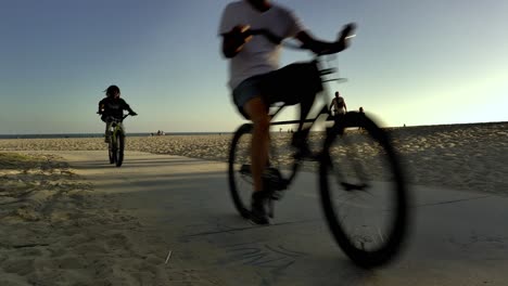 People-riding-bikes-along-Venice-Beach-Boardwalk-during-sunset-golden-hour,-in-Los-Angeles,-Califonia,-USA---Low-Angle