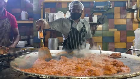 Indian-Cook-Mixing-Sauce-In-A-Large-Wok-With-A-Flat-Laddle-In-The-Street-Of-New-Delhi,-India