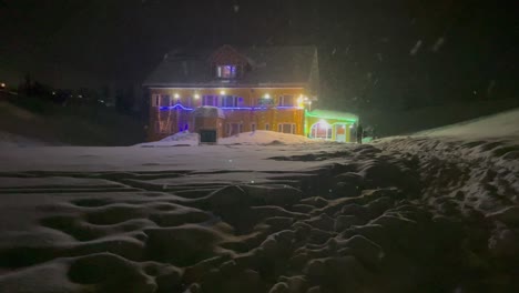 People-Walking-At-Night-On-Snowy-Landscape-Outside-The-House-Adorned-With-Christmas-Lights-Amidst-Snowfall-In-Winter-In-Gulmarg,-India