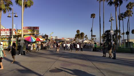 People-walking-between-shops-and-the-beach-at-the-Venice-Boardwalk,-during-golden-hour,-in-Los-Angeles,-California,-USA---Static-shot