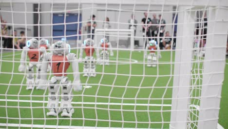 Follow-Shot-Behind-Football-Goal-Netting-With-Nao-Football-Robots-Standing-On-Pitch