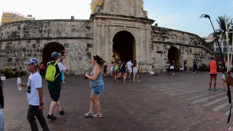 Men-and-women,-tourists-are-walking-and-taking-pictures-with-their-cameras-near-an-old-wall,-arc-and-clock-of-a-plaza-in-the-old-town-of-Cartagena-de-Indias,-Colombia