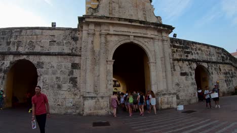 A-group-of-tourists-and-a-man-with-a-red-shirt-are-walking-near-an-old-arc-or-door-of-stones-that-enters-a-plaza-of-the-old-town-of-Cartagena-de-Indias,-Colombia