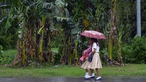 Dos-Jóvenes-Alumnas-Filipinas-Caminan-Juntas-A-Casa-Bajo-La-Lluvia-A-Lo-Largo-De-Una-Carretera-Bordeada-De-Palmeras