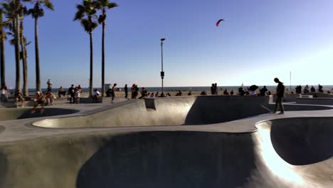 People-skating-at-the-Venice-Beach-Skate-Park-with-ocean-and-mountains-in-background,-on-a-sunny-day,-in-Los-Angeles,-California,-USA---Pan-shot