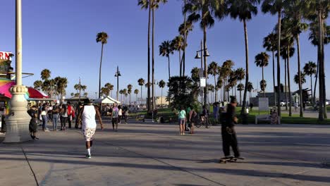 People-walking-and-riding-skateboard-around-the-Venice-Boardwalk-wearing-masks,-during-golden-hour,-in-Los-Angeles-during-Covid-19-pandemic---handheld-static-shot