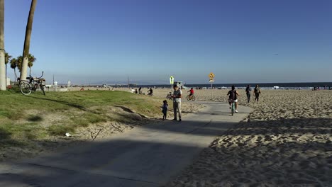 Menschen,-Die-Fahrrad-Fahren-Und-Auf-Der-Strandpromenade-Von-Venedig-Mit-Dem-Meer-Im-Hintergrund-In-Los-Angeles,-Kalifornien,-Spazieren-Gehen-–-Handaufnahme