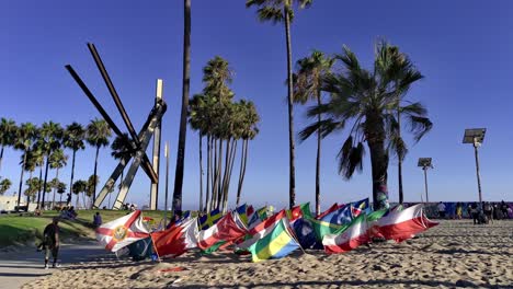 Multiple-multi-country-Flags-waving-in-a-the-wind,-while-people-walk-by,-at-the-Venice-beach-Boardwalk,-during-the-day,-in-LA,-California,-USA---static-shot-slow-motion