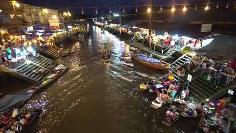 Nacht-Im-Geschäftigen-Thailändischen-Schwimmenden-Markt,-Bootslebensmittel--Und-Obstverkäufer-Und-Restaurants-Am-Flussufer,-Panorama