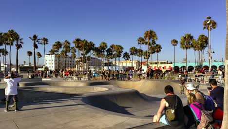People-skating-and-sitting-around-at-the-Venice-Beach-Skate-Park-with-ocean-and-mountains-in-background,-on-a-sunny-day,-in-Los-Angeles,-California,-USA---handheld-shot