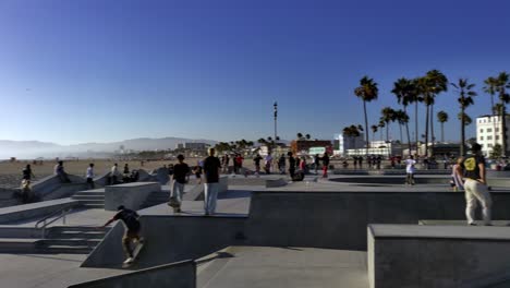 People-skating-at-the-Venice-Beach-Skate-Park-with-beach-and-mountains-in-background,-on-a-sunny-day,-in-Los-Angeles,-California,-USA---Pan-shot
