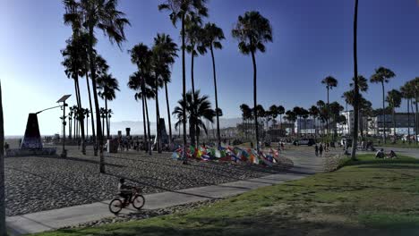 People-walking-and-riding-bikes-along-Venice-Beqch-Boardwalk-with-skatepark-in-backgound-at-sunset,-golden-hour,-in-Los-Angeles,-CA