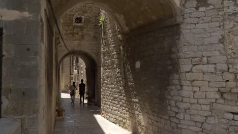 Two-people,-a-woman-and-a-man-walking-through-the-old-town-alleys-of-Sibenik-under-a-tunnel-in-a-narrow-alley-protected-by-shadow-in-a-hot-sunny-summer-day