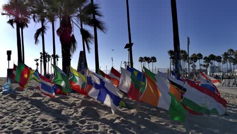 Multiple-multi-country-Flags-waving-in-a-the-wind,-while-people-walk-by,-at-the-Venice-beach-Boardwalk,-during-golden-hour,-in-LA,-California,-USA---static-shot