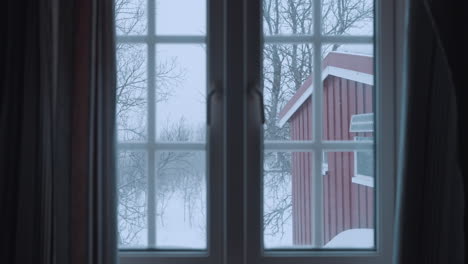 Gemütlicher-Blick-Aus-Einem-Norwegischen-Hüttenfenster,-Der-Eine-Ruhige-Winterwaldszene-Mit-Sanft-Fallendem-Schnee-Und-Bezaubernden-Roten-Hütten-Inmitten-Schneebedeckter-Bäume-Einfängt