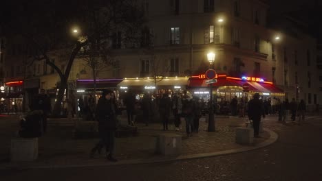 People-walking-over-square-in-Paris,-during-night-time,-street-lights-and-restaurants-illuminate-the-street
