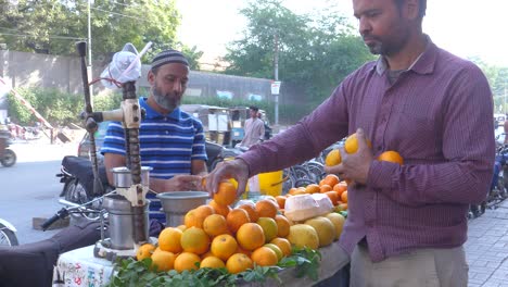 Backward-closeup-shot-of-two-men-selling-orange-juices-during-daytime-at-a-roadside-stall-in-Saddar-Bazar-Street-of-Karachi,-Pakistan