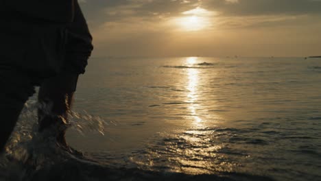 Nahaufnahme-Der-Füße-Eines-Mannes,-Der-Bei-Sonnenaufgang-In-Zeitlupe-Am-Strand-Im-Wasser-Läuft
