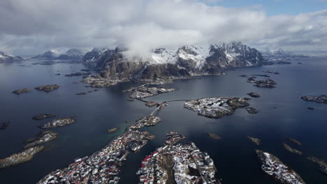 Drone-footage-over-Henningsvær,-showcasing-the-stunning-contrast-of-snowy-mountains,-vibrant-blue-waters,-and-scattered-islands-of-the-Lofoten-archipelago-under-a-cloud-adorned-sky