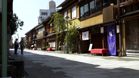 A-daytime-scene-captures-the-Nishi-Chaya-District-in-Kanazawa,-Japan,-featuring-people-strolling-along-the-street-amidst-passing-car-traffic-and-showcasing-typical-Japanese-architecture