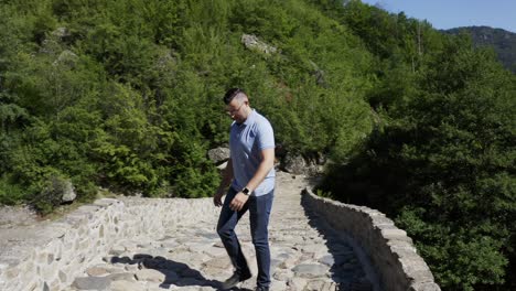 A-shot-of-a-tourist-looking-down-the-Arda-River-and-walking-down-the-Devil's-Bridge-on-a-cobblestone-pathway,-located-in-the-town-of-Ardino-at-the-foot-of-Rhodope-Mountains-in-Bulgaria