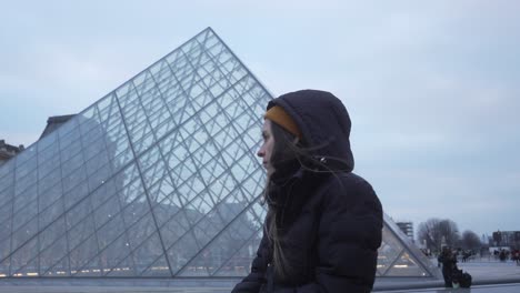 Female-tourist-portrait,-relaxing-at-the-Louvre-Pyramid-during-cloudy-winter-day