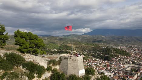 Filmische-Drohnenaufnahme-Der-Albanischen-Nationalflagge,-Die-An-Einem-Historischen-Gebäude-Mit-Blick-Auf-Die-Stadt-Barat-Und-Einer-Malerischen-Landschaft-Unter-Einem-Bewölkten-Himmel-In-Europa-Weht