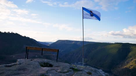 Galicia-flag-blows-strong-in-wind-overlooking-Ribeira-Sacra-and-canyon-overlook-with-empty-bench