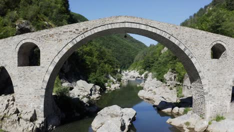 Retreating-drone-shot-over-the-Arda-River-and-going-through-the-main-arch-of-the-Devil's-Bridge-and-revealing-the-beautiful-landscape-at-the-foot-of-the-Rhodope-Mountains-in-Bulgaria