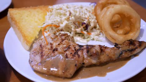 Close-up-of-a-platter-of-chicken-steak-in-barbecue-sauce,-served-alongside-some-onion-rings,-a-coleslaw-salad,-and-a-slice-of-buttered-toasted-bread