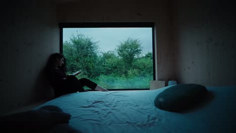 Woman-reading-a-book-during-dusk-by-window-in-a-cabin-in-nature-wide-shot