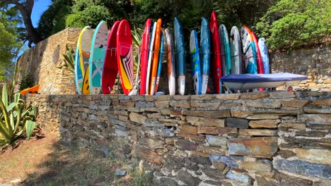 Colourful-stand-up-puddle-board-collection-in-a-tropical-villa-by-the-beach-in-South-of-France-with-green-trees-and-blue-sky,-fun-sunny-vacation-sport-holiday-activity,-4K-shot
