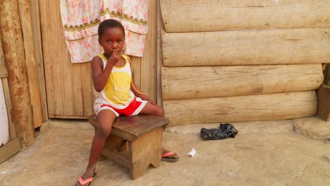 little-cute-black-African-girl-sitting-on-a-wooden-chair-while-eating-candy-in-her-small-remote-rural-village-of-Africa