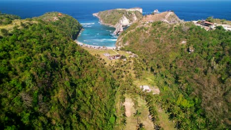 Tropical-Palms-Valley-With-Few-Indonesian-Houses-Leading-to-Atuh-Beach-and-Batupadasan-Island-in-Nusa-Penida,-Bali,-Indonesia---Aerial-view