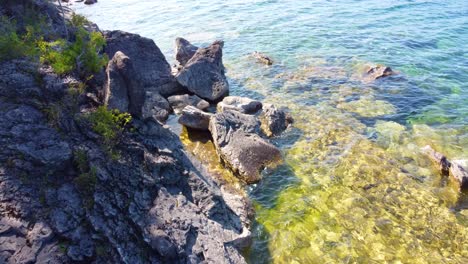 Aerial-drone-shot-descending-down-over-rocks-revealing-the-crystal-clear-waters-which-surround-the-beautiful-coastline-of-Georgian-Bay,-Ontario,-Canada