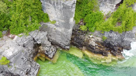 Aerial-top-down-view-of-the-ocean-waves-washing-up-against-a-rocky-cliff-along-the-coastline-of-Georgian-Bay,-Ontario,-Canada