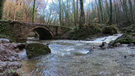 Fluss-Fließt-Aus-Einem-Kleinen-Waldwasserfall-Unter-Einer-Rustikalen-Steinfußgängerbrücke-Im-Herbstwald-In-Zeitlupe
