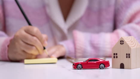 Close-up-of-a-hand-holding-a-pencil-and-writing-something-on-a-yellow-paper,-behind-a-red-toy-car-that-signifies-car-loan-financing