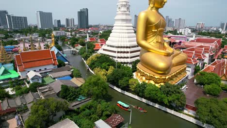 Chao-Phraya-River-Cruise-Boats-Passing-Big-Golden-Buddha-Statue-Wat-Paknam-Bhasicharoen-in-Phasi-Charoen-District-in-Bangkok,-Thailand---aerial-view